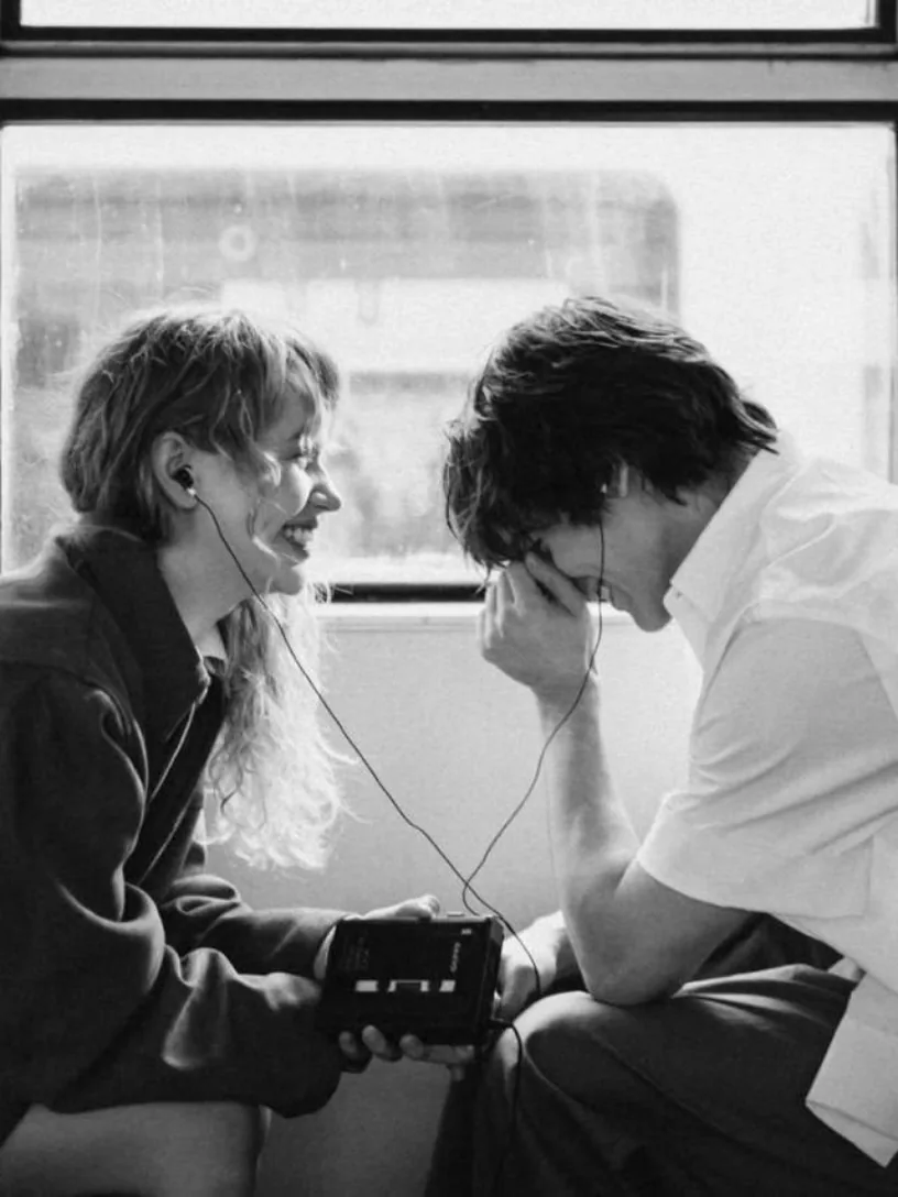 A black-and-white photograph of a couple leaning together over a shared device on a train, from the MutualClause campaign